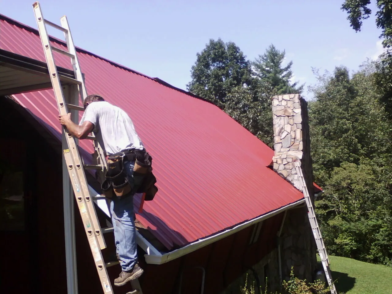 Roofer on a ladder beside a red metal roof and stone chimney — Soffit & Fascia Repair by Roofing Brothers in Umatilla